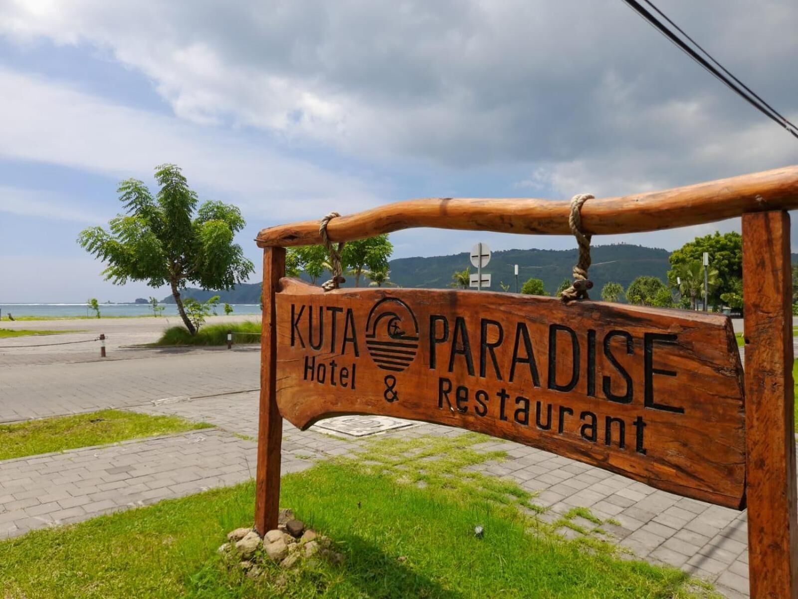Une enseigne en bois indiquant l’hôtel-restaurant Kuta Paradise, avec une vue sur la plage et les montagnes en arrière-plan.