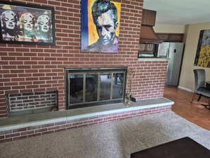 Una sala de estar con chimenea de ladrillo, un cuadro grande encima y una cocina al fondo.