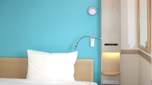 Une chambre d’hôtel avec un lit, un thermostat fixé au mur et une petite étagère avec un livre.