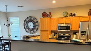 A modern kitchen with wooden cabinets, a stainless steel refrigerator, a microwave, and a large clock on the wall. There are three black barstools and a patterned rug on the floor. - Oasis at Johnson Ranch (Queen Creek)