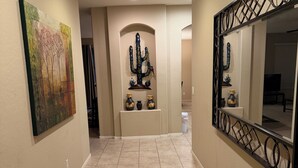 A hallway with beige walls, a tiled floor, a cactus decoration, and a large mirror on the right. - Oasis at Johnson Ranch (Queen Creek)