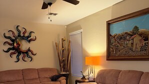 A living room with two brown sofas, a wooden coffee table, a ceiling fan, a decorative sun sculpture, a lamp, and a framed painting. - Oasis at Johnson Ranch (Queen Creek)