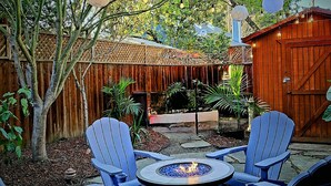 A patio area with a fire pit table surrounded by blue Adirondack chairs, a wooden fence, and string lights.