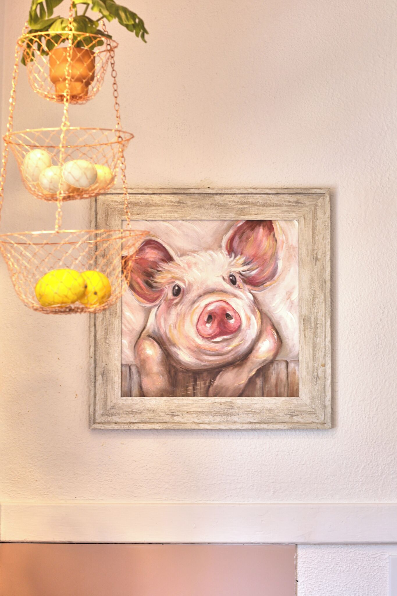 A wall with a framed painting of a pig and hanging baskets with fruits.