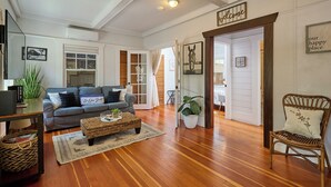 A spacious living room with a grey sofa, a coffee table, and a wicker chair. There is a wooden floor, a large window, and a door leading to another room.