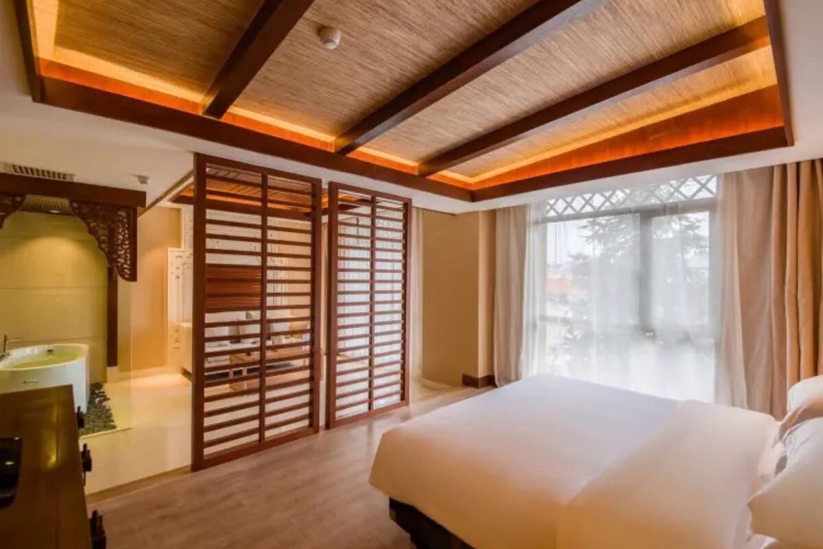 A hotel room with a large bed, wooden ceiling beams, a window with curtains, and a bathroom with a sink and mirror.