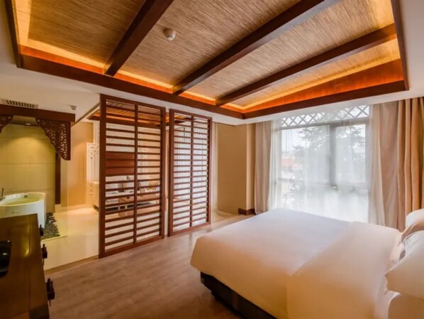 A hotel room with a large bed, wooden ceiling beams, a window with curtains, and a bathroom with a sink and mirror.