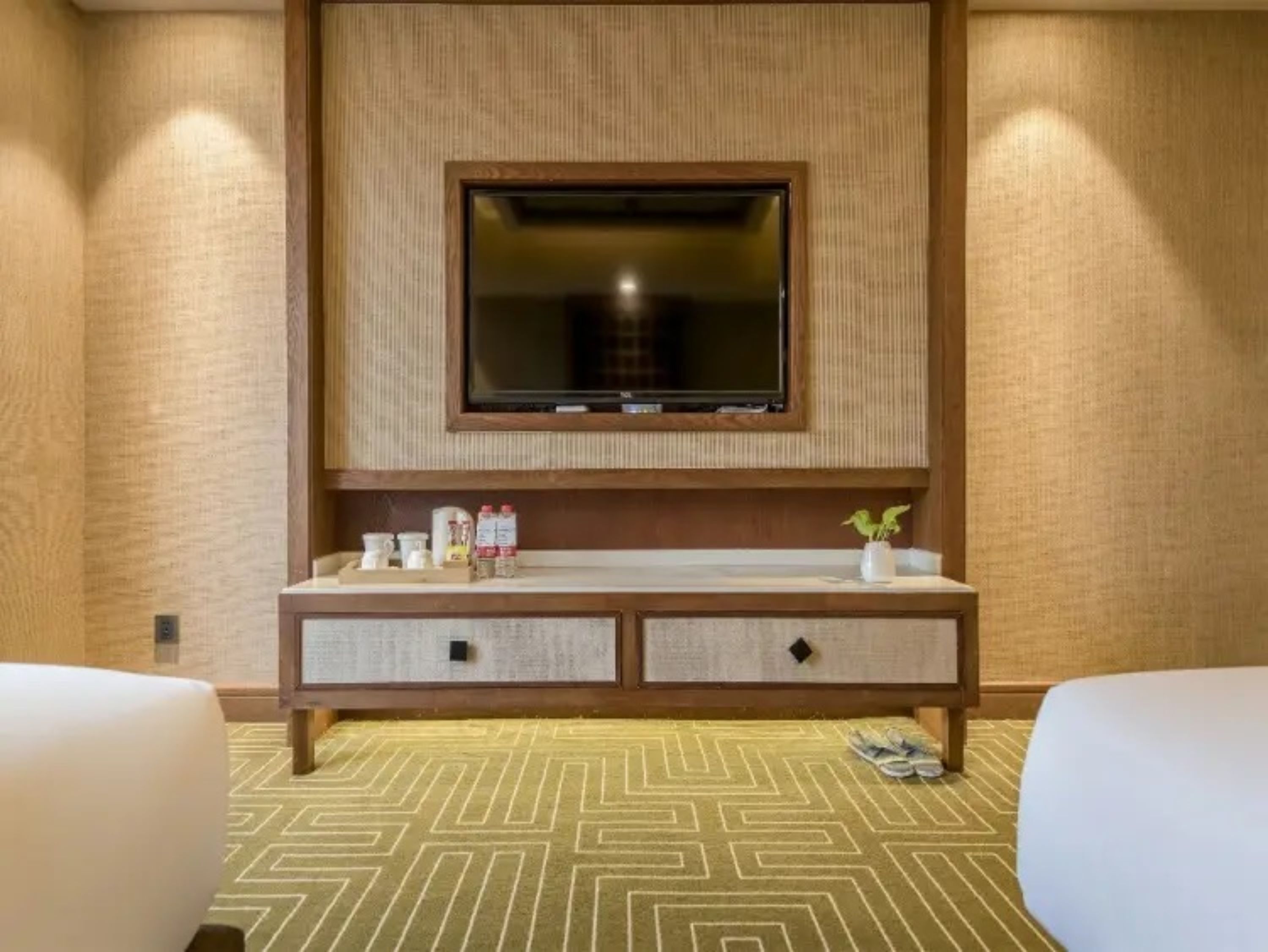 A hotel room with a large flat-screen TV mounted on a wooden panel wall, a small bedside table with a vase of flowers, and a patterned carpet.