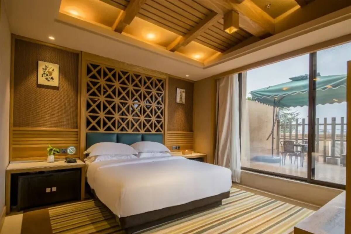 A hotel room with a large bed, wooden headboard, a decorative wall panel, a bedside table, and a view of the outdoors through a large window.