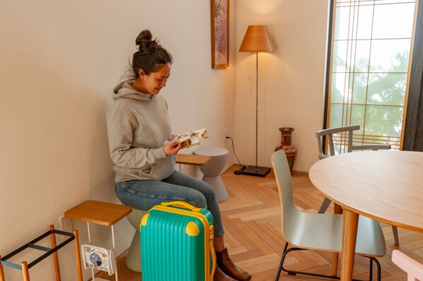A person sitting on a bench, reading a book in a room with a round table, chairs, a lamp, and a window. - plat hostel keikyu haneda home (Tokyo)