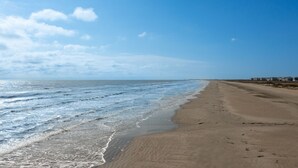 Una amplia playa de arena con olas suaves, cielo despejado y una costa lejana con edificios.