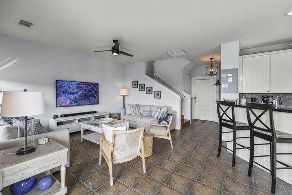 Amplia sala de estar con sofá, mesa de centro, sillas, televisor y cocina con armarios blancos y barra.