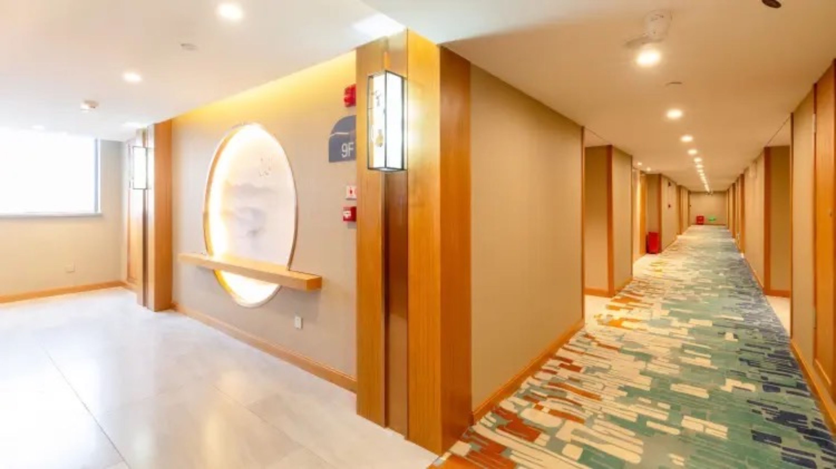 Corredor de hotel com carpete estampado, paredes de madeira e luminária fixada na parede.