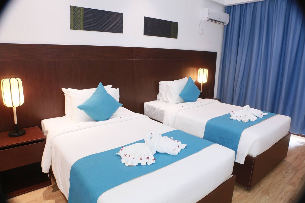 Ein Hotelzimmer mit zwei Betten, jeweils mit weißer Bettwäsche und blauen Akzenten, einem Holz-Kopfende und einem blauen Vorhang.