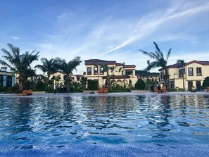 A swimming pool with lounge chairs and palm trees, surrounded by buildings with a Mediterranean architectural style. - Sealong Bay ZhongQi Conifer Hotel (Prey Nob)