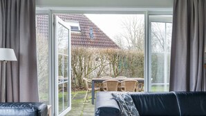 A modern living room with a black leather sofa, a table with wicker chairs, and a view of a garden and house through large glass doors. - Bungalowpark It Wiid by Interhome (Eernewoude)
