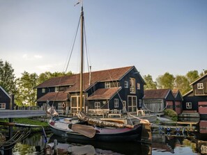 Une maison traditionnelle en bois, dotée d’un toit à pignon très incliné, d’un quai avec une barque à rames et d’un ciel dégagé.
