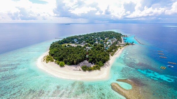 An aerial view of a tropical island with a beach, clear turquoise waters, and a small settlement with buildings and a dock. - Kuri Beach View Inn (Omadhoo)