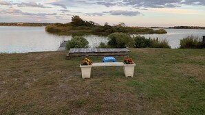 Vista de un lago con un muelle de madera que se adentra en el agua, un banco con flores y una zona con césped.
