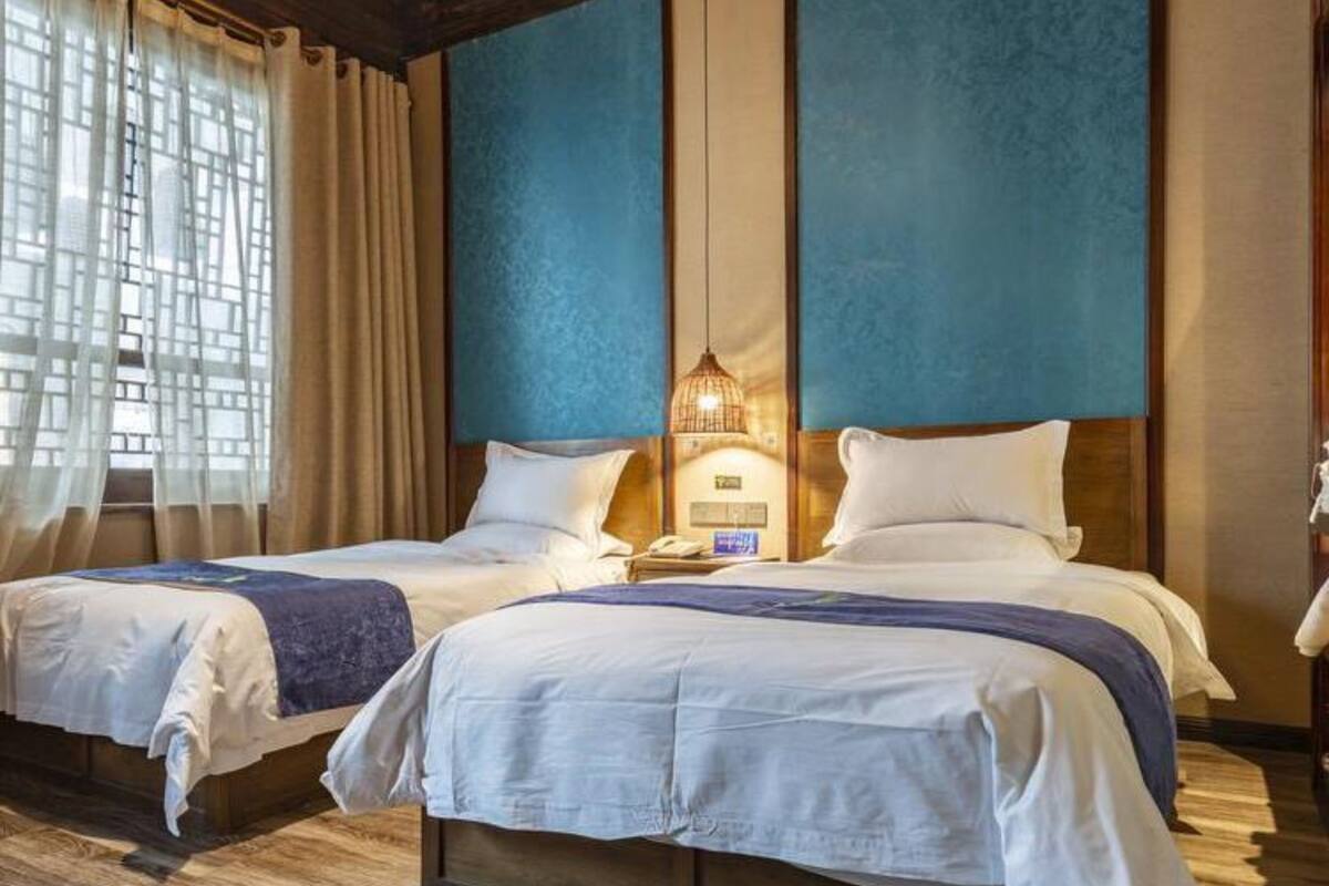 A hotel room with two beds, a wooden floor, a blue accent wall, and a window with curtains.