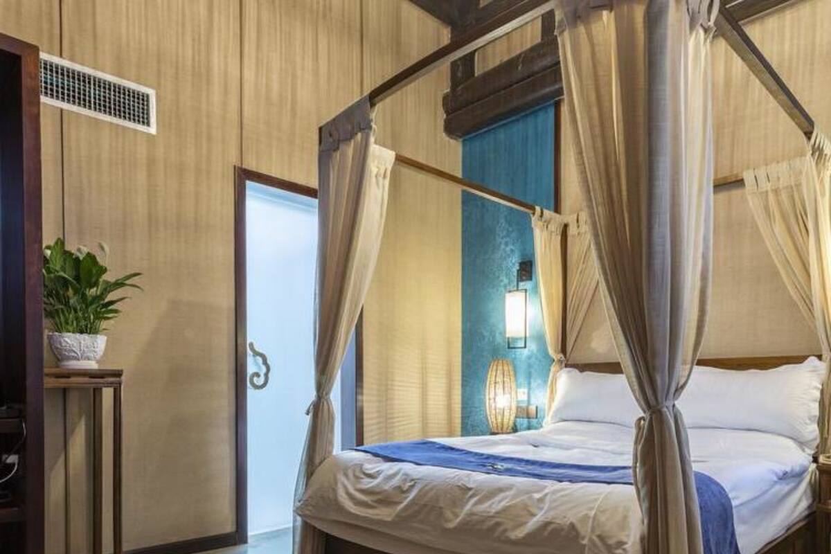A four-poster bed with white linens and a blue throw blanket, a bedside lamp, and a potted plant on a wooden nightstand.