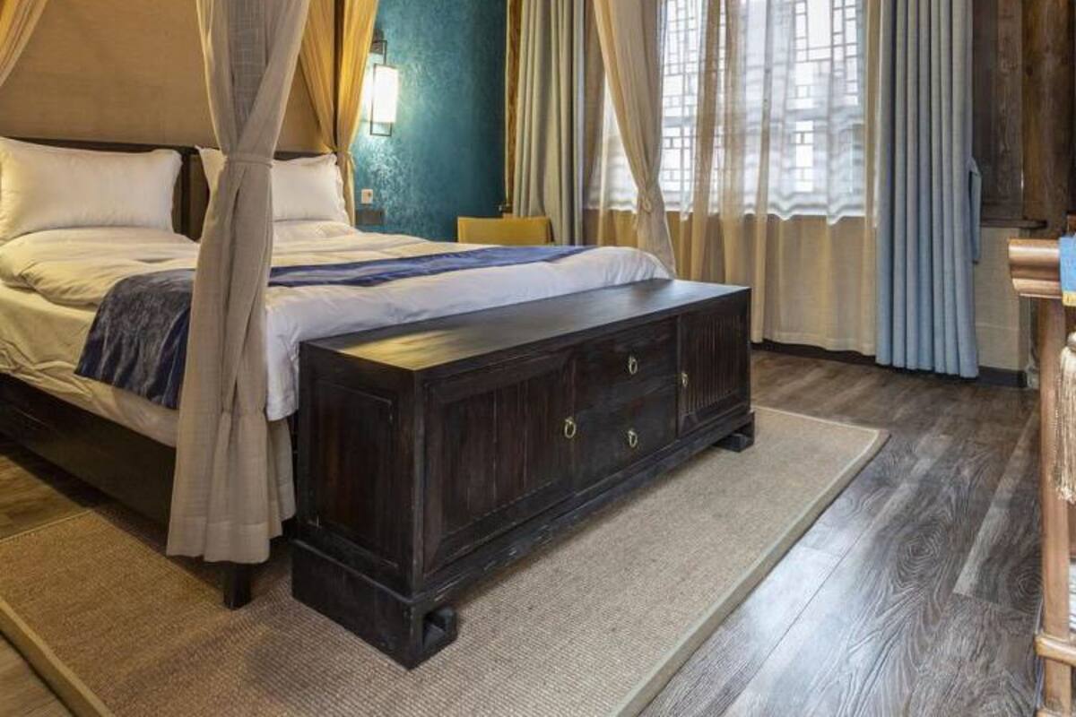 A four-poster bed with a dark wooden bench, a large window with curtains, and a wooden ceiling.