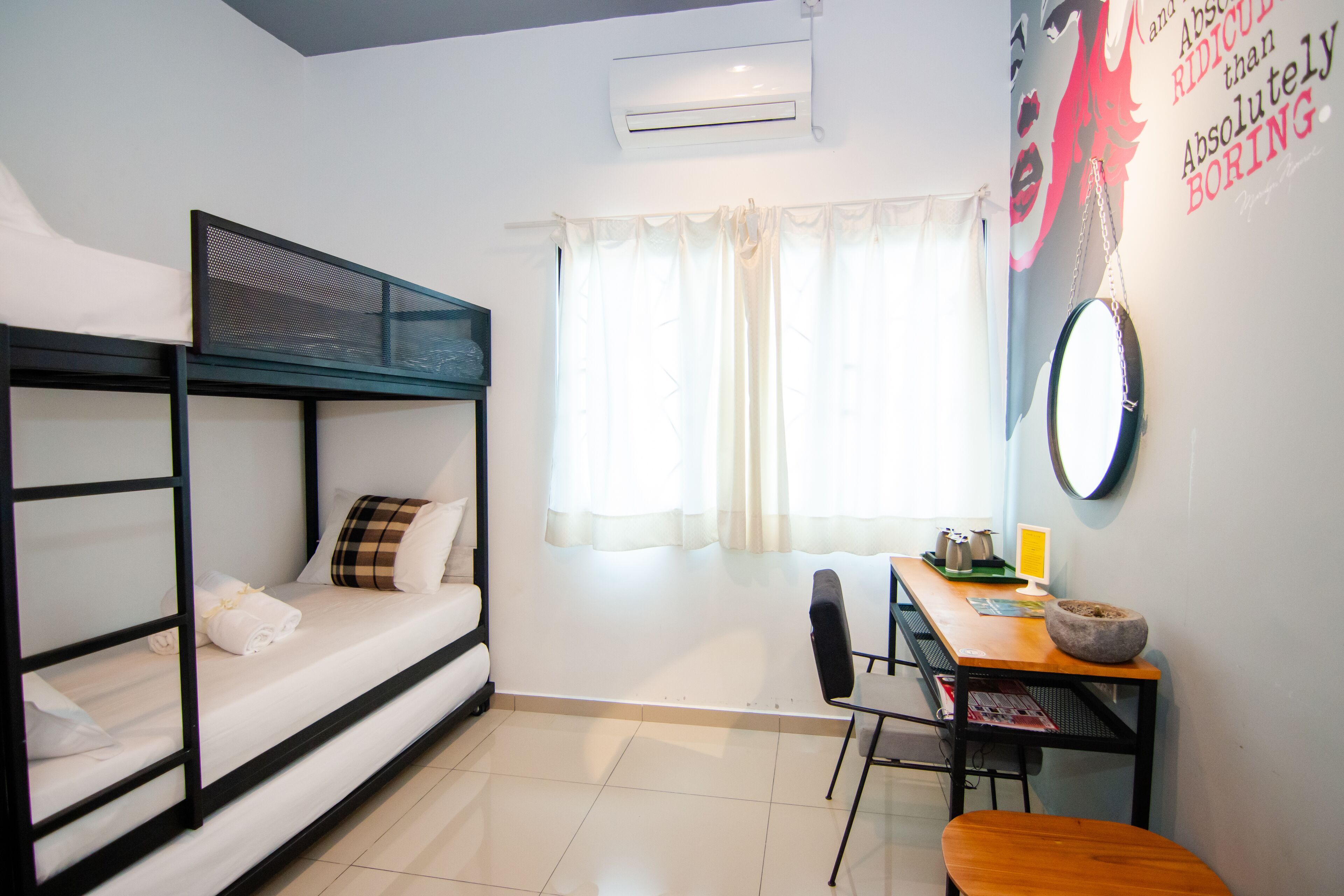 A dorm-style room with bunk beds, a desk, a chair, a table, and a window with curtains.