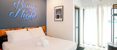 A neatly made bed with white linens and a towel, a wall sign reading "Bring on the Night", and a framed picture on the wall.