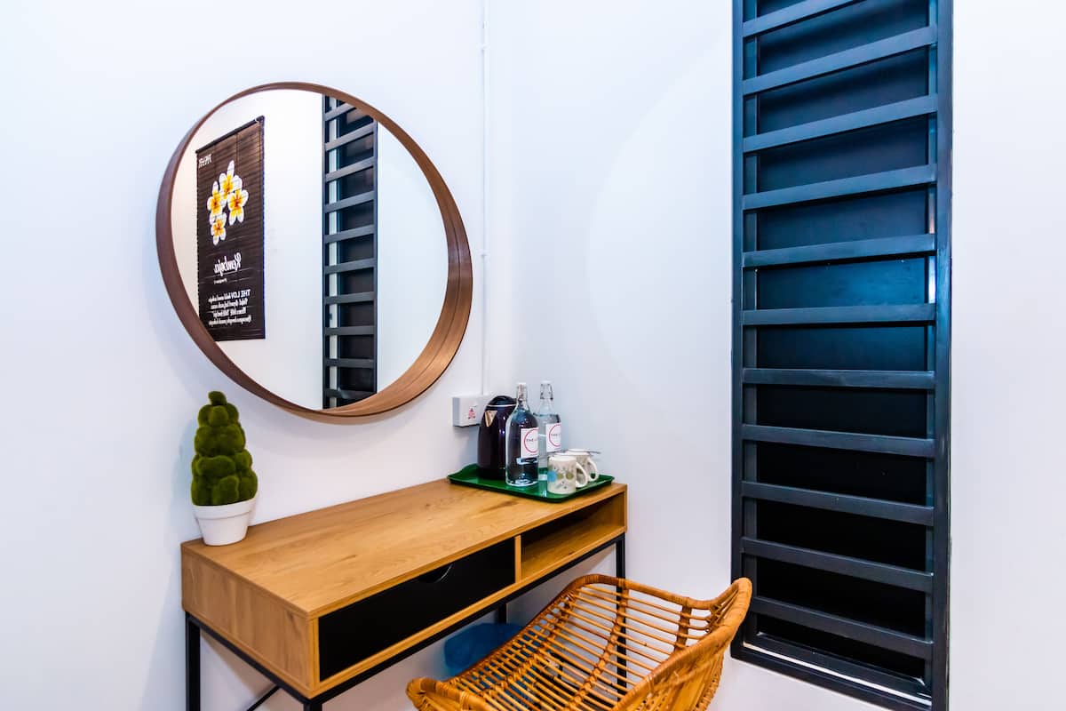 A modern room with a wooden console table, a round mirror, a small potted plant, a wicker chair, and a window with black blinds.