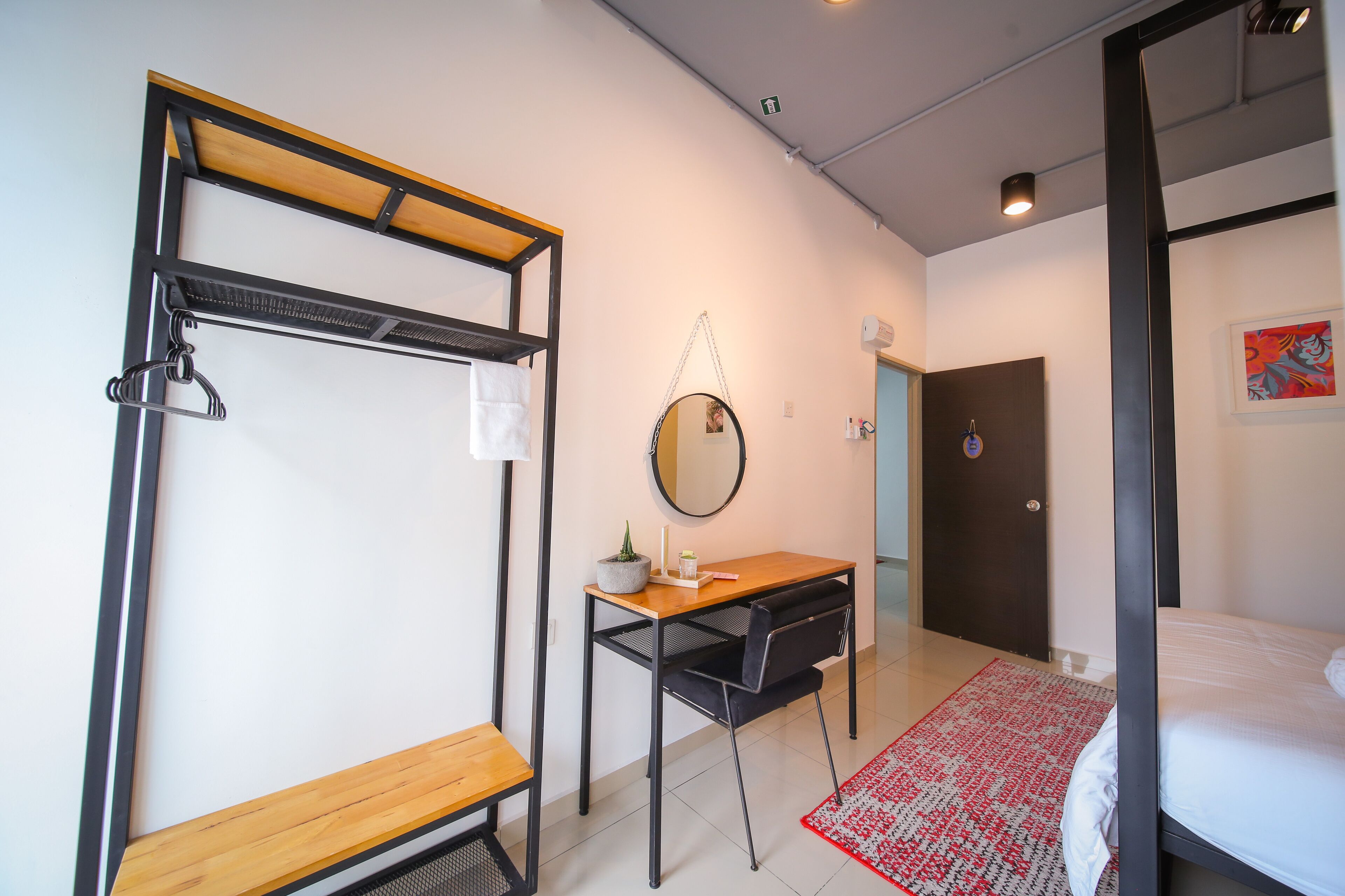 A compact hotel room with a bed, a desk with a chair, a hanging coat rack, a small table with a plant, and a mirror.