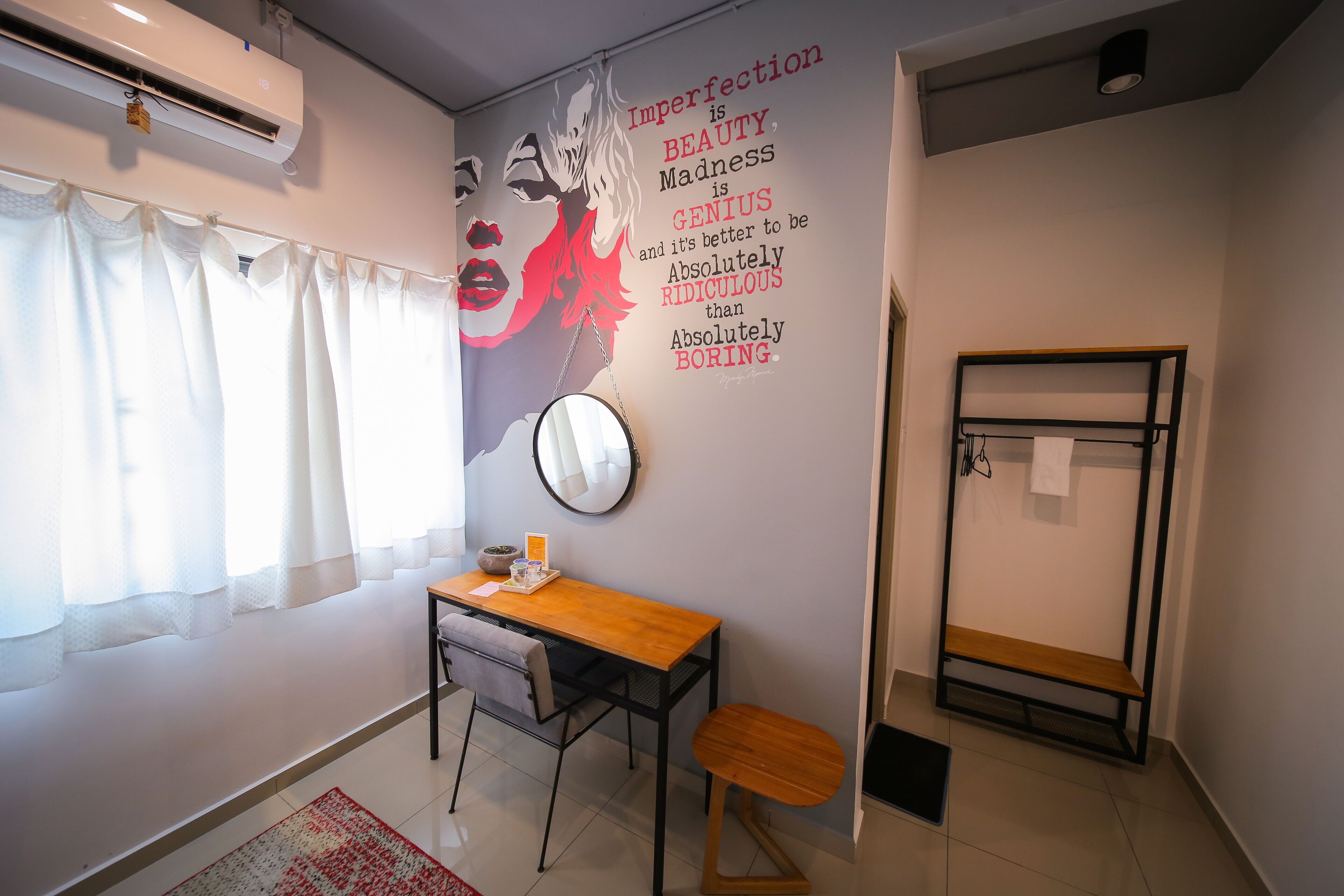 Una habitación con escritorio, silla y espejo en la pared. Se ve una ventana con cortinas, un radiador y un banco pegado a la pared.