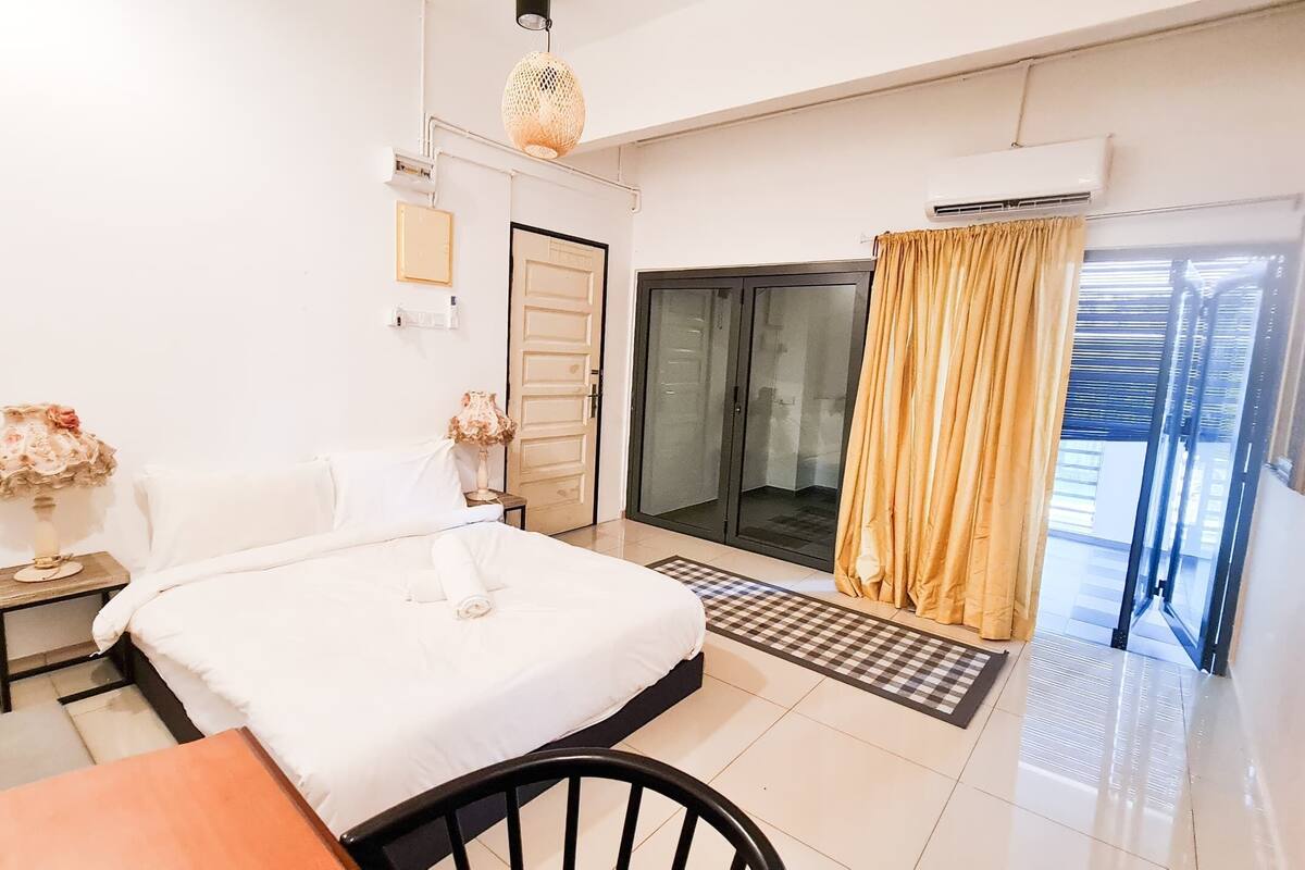 A bedroom with a bed, a nightstand, a lamp, a window with curtains, and an air conditioning unit.