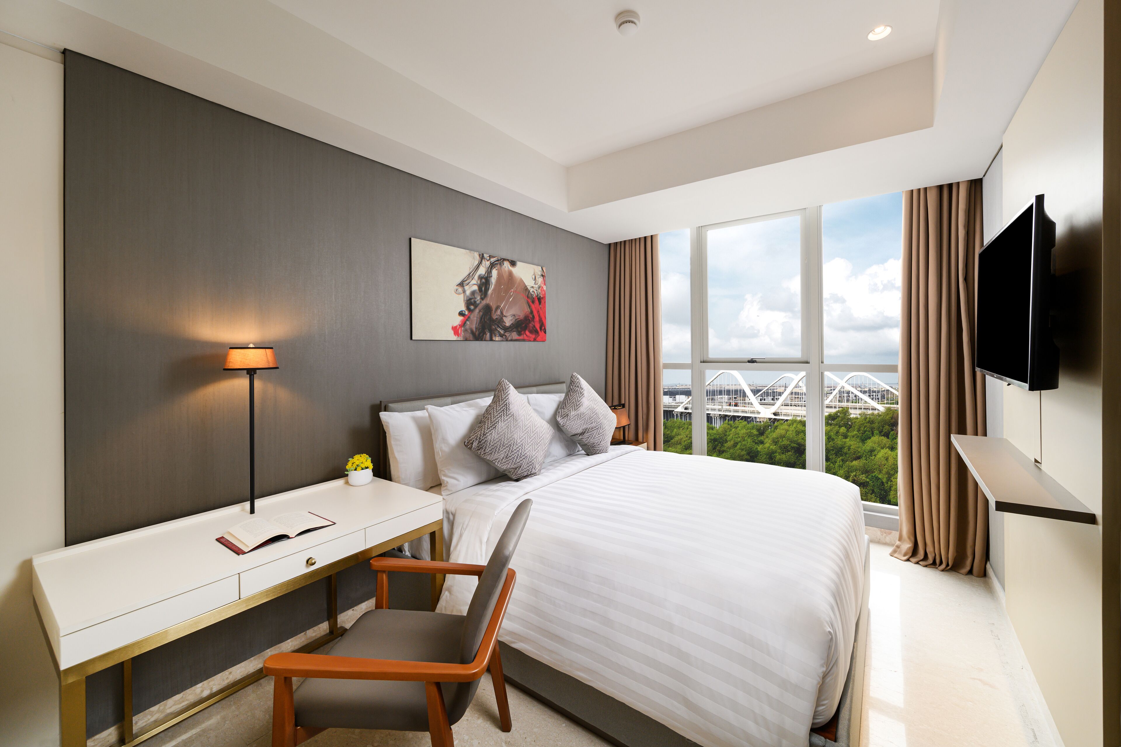 Habitación de hotel moderna con una cama grande, un escritorio con lámpara, una silla, televisor de pantalla plana y un ventanal con vistas a la ciudad.