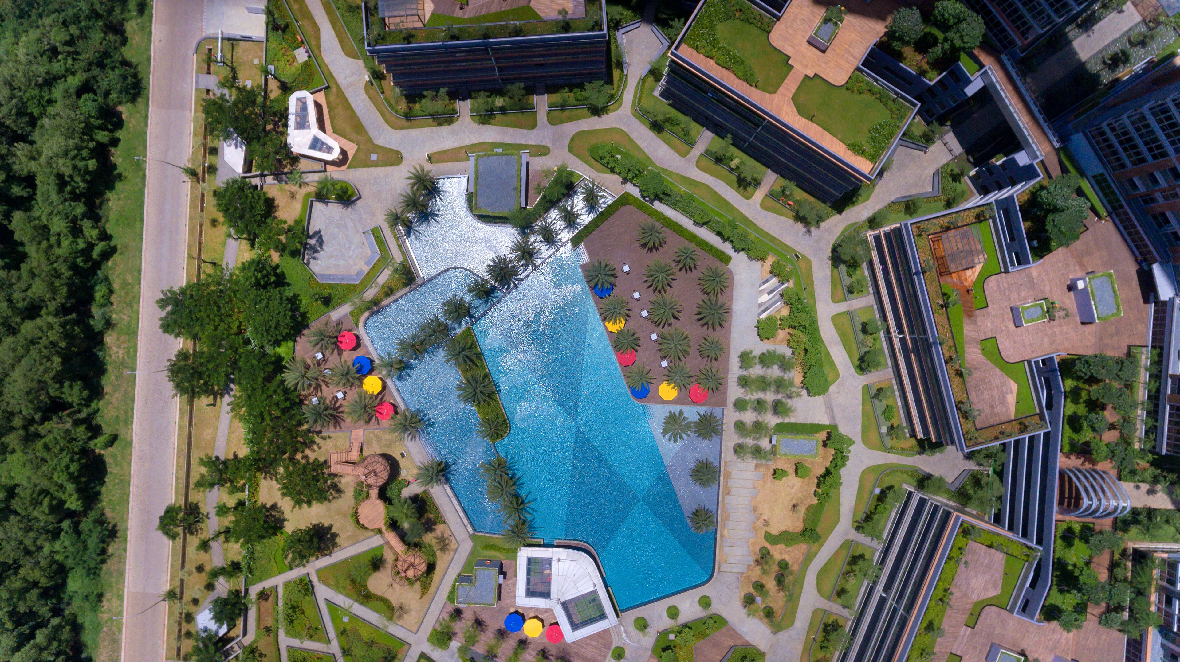 An aerial view of a modern hotel with a central swimming pool, surrounded by landscaped gardens and palm trees.