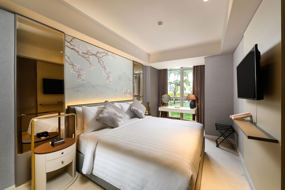 A modern hotel room with a large bed, a flat-screen TV mounted on the wall, a bedside table with a lamp, and a view of greenery outside.