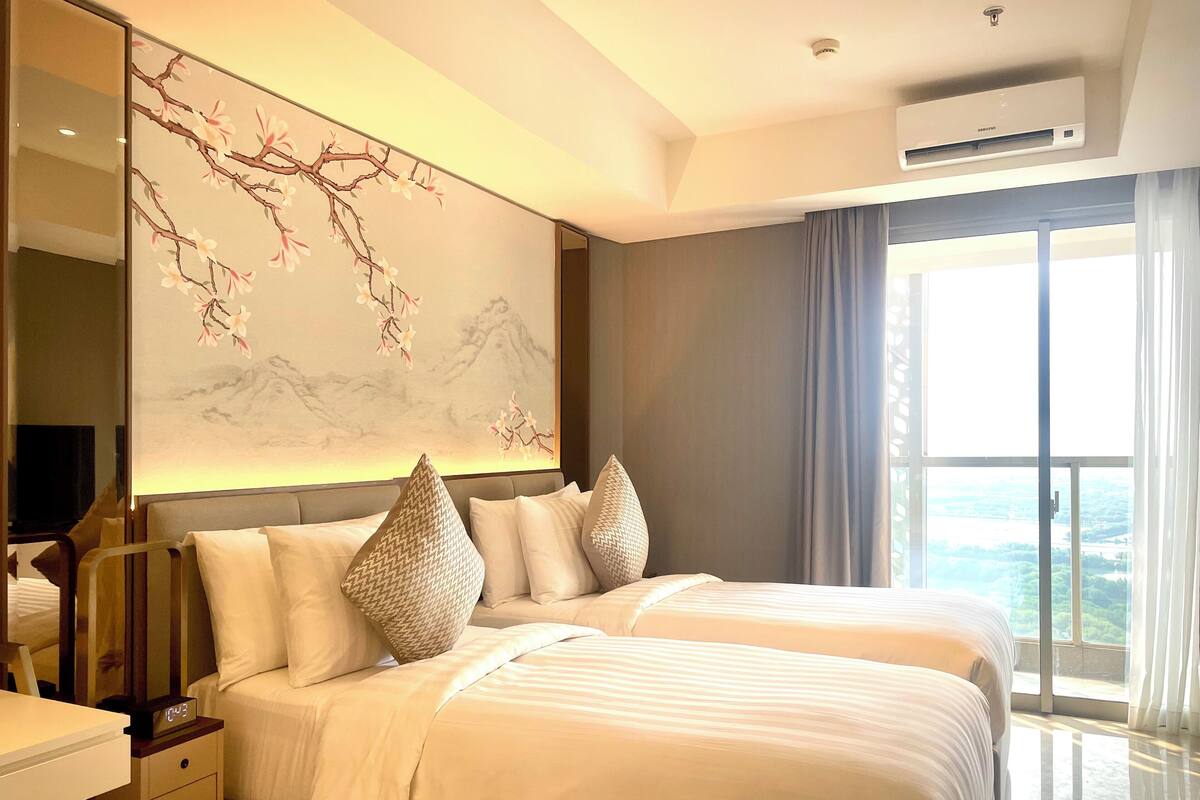 A hotel room with two beds, a large window with a view, a wall-mounted air conditioner, and a bedside table with a clock.