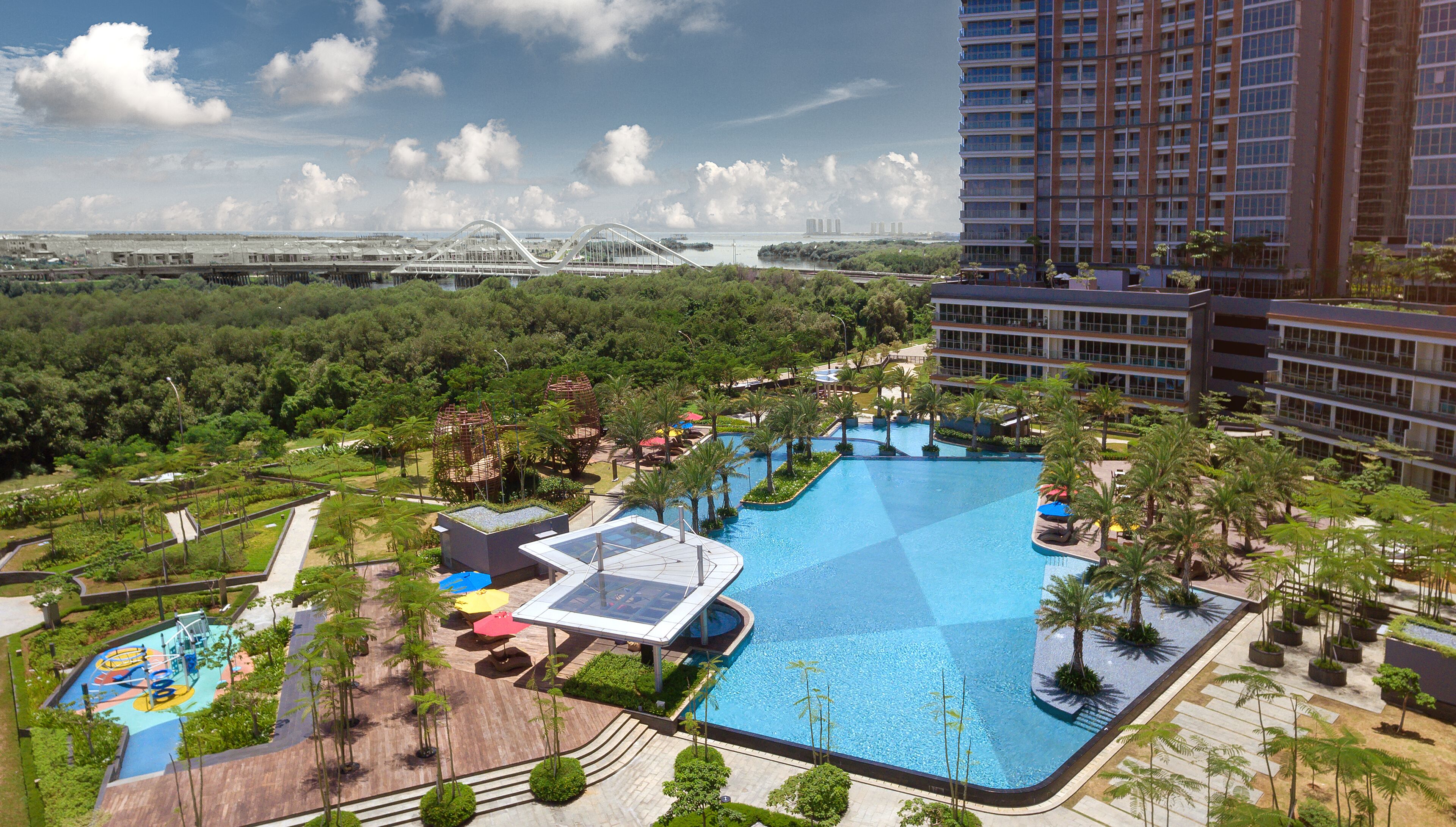A large swimming pool with a sun deck, surrounded by palm trees and a view of a bridge and buildings in the background.