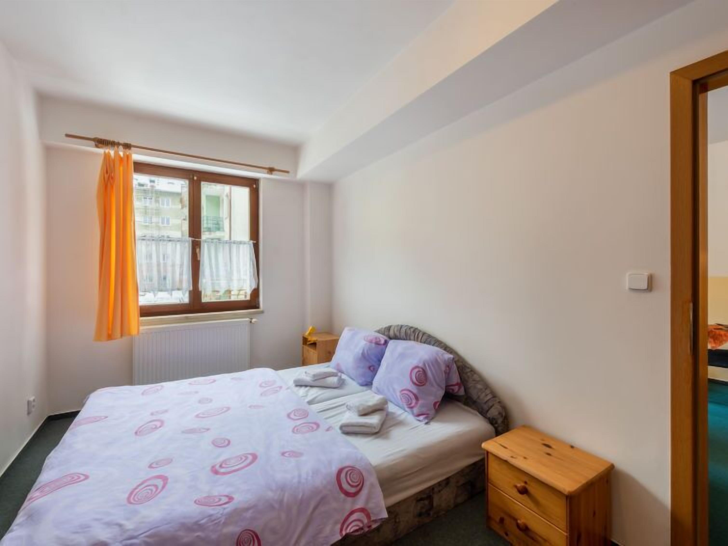Une chambre à coucher avec un lit, une table de chevet en bois, une fenêtre avec des rideaux et un radiateur.