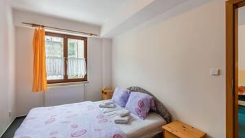 Une chambre à coucher avec un lit, une table de chevet en bois, une fenêtre avec des rideaux et un radiateur.