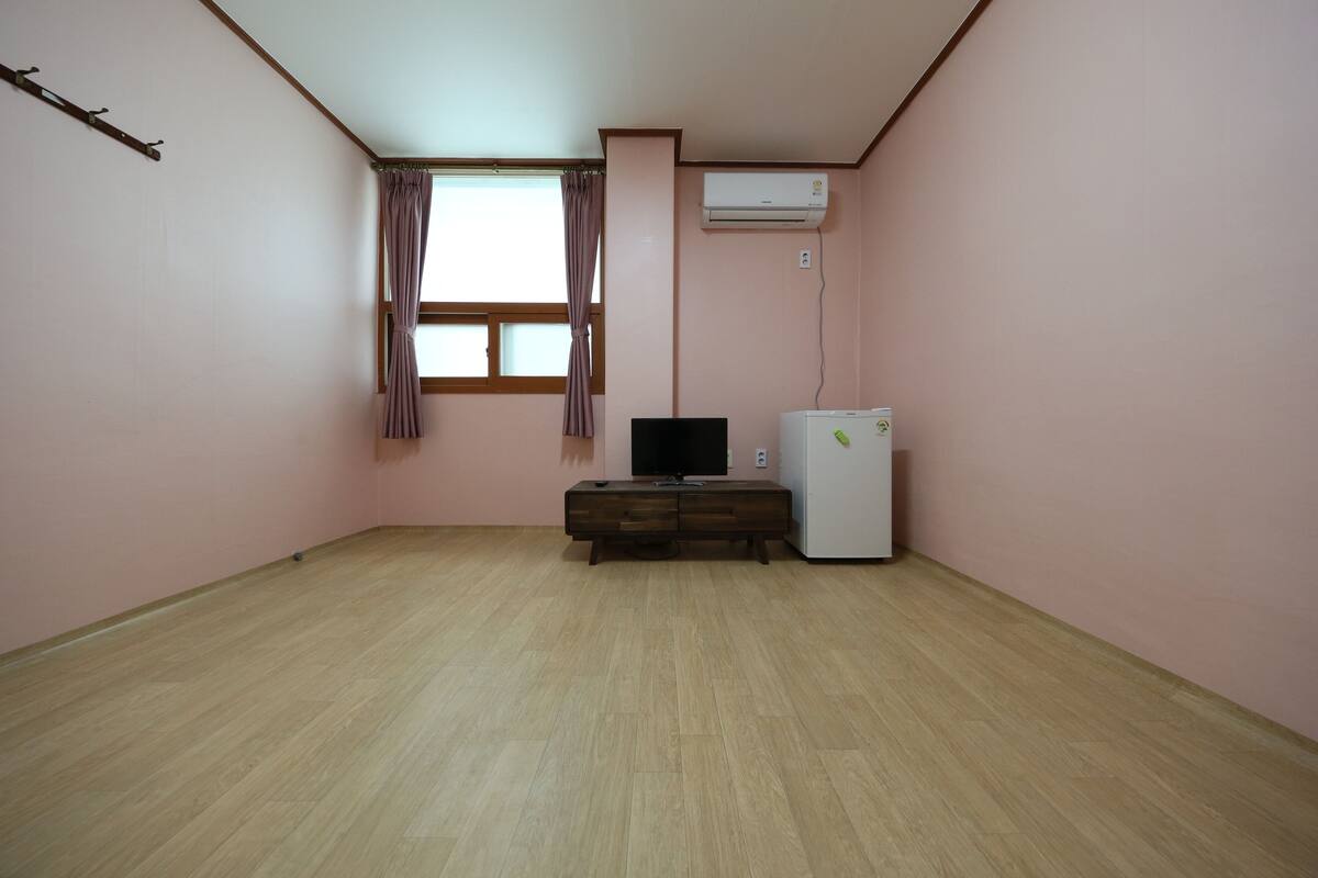 Una habitación con paredes rosas, piso de madera, televisor de pantalla plana, un refrigerador pequeño y una ventana con cortinas.