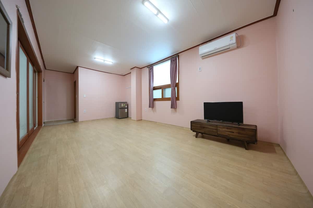 Una habitación amplia con piso de madera, un televisor de pantalla plana sobre un mueble de madera, aire acondicionado en la pared y una ventana con cortinas.