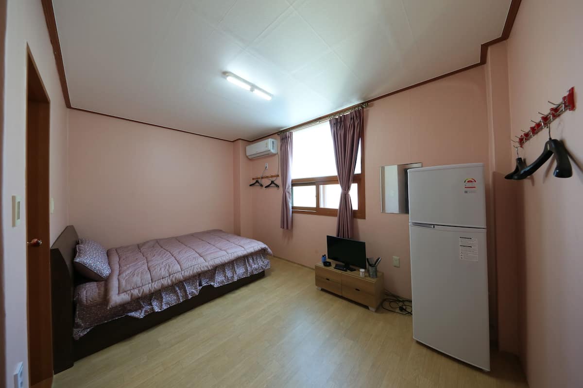 Un dormitorio pequeño y bien iluminado con una cama individual, televisor, refrigerador y ventana con cortinas.