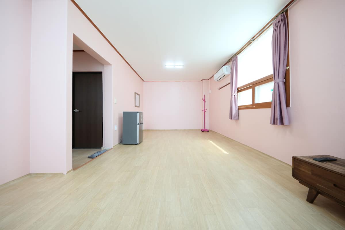 Una habitación amplia con piso de madera, una puerta, un refrigerador pequeño, una ventana con cortinas y un aparador de madera.