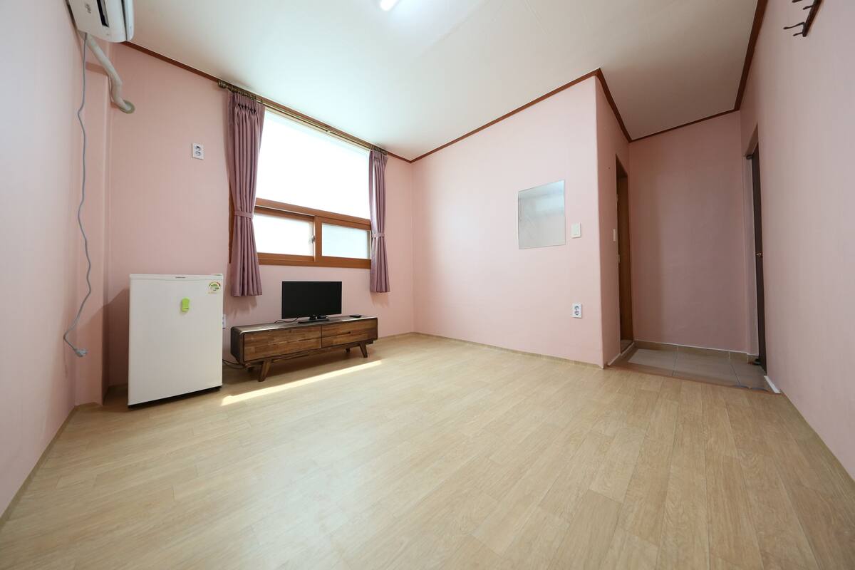 Una habitación con paredes rosas, suelo de madera, un refrigerador blanco, un armario de madera, una televisión y una ventana con cortinas.