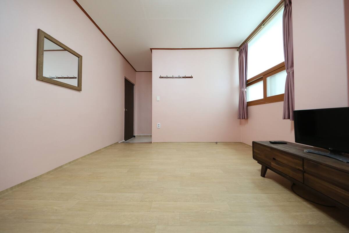 Una habitación con piso de madera, un televisor sobre un mueble de madera, un espejo en la pared y una ventana con cortinas.