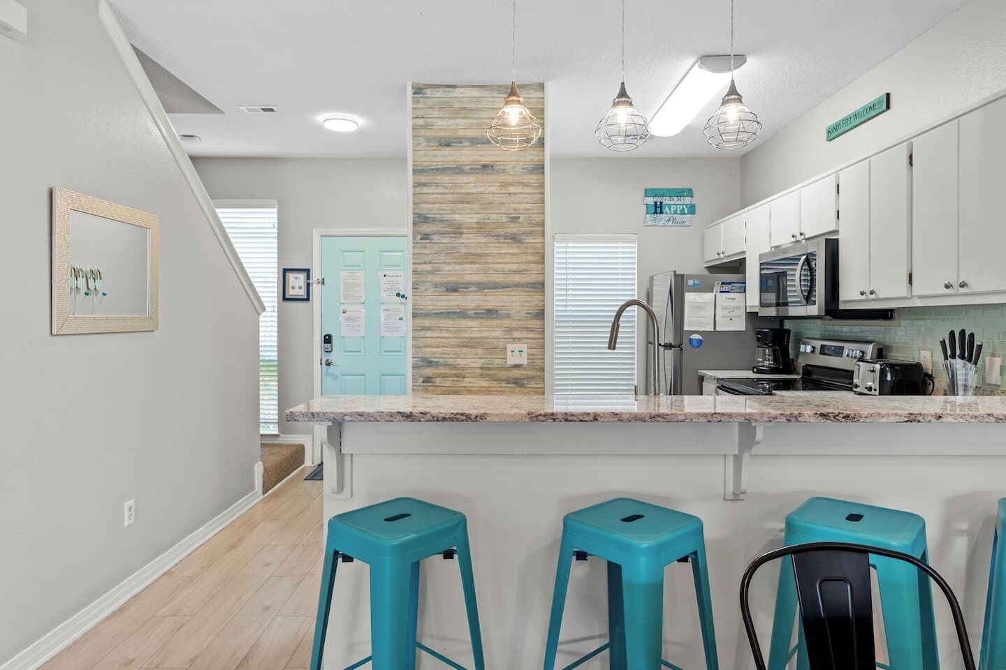Een moderne keuken met witte kasten, een granieten aanrechtblad en drie turquoise barstoelen.
