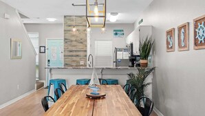 A modern kitchen with a wooden table and blue chairs, a hanging light fixture, and decorative wall art. - Gem Beach House Steps to Beach&Pool+Opt Golf Cart (Miramar Beach)