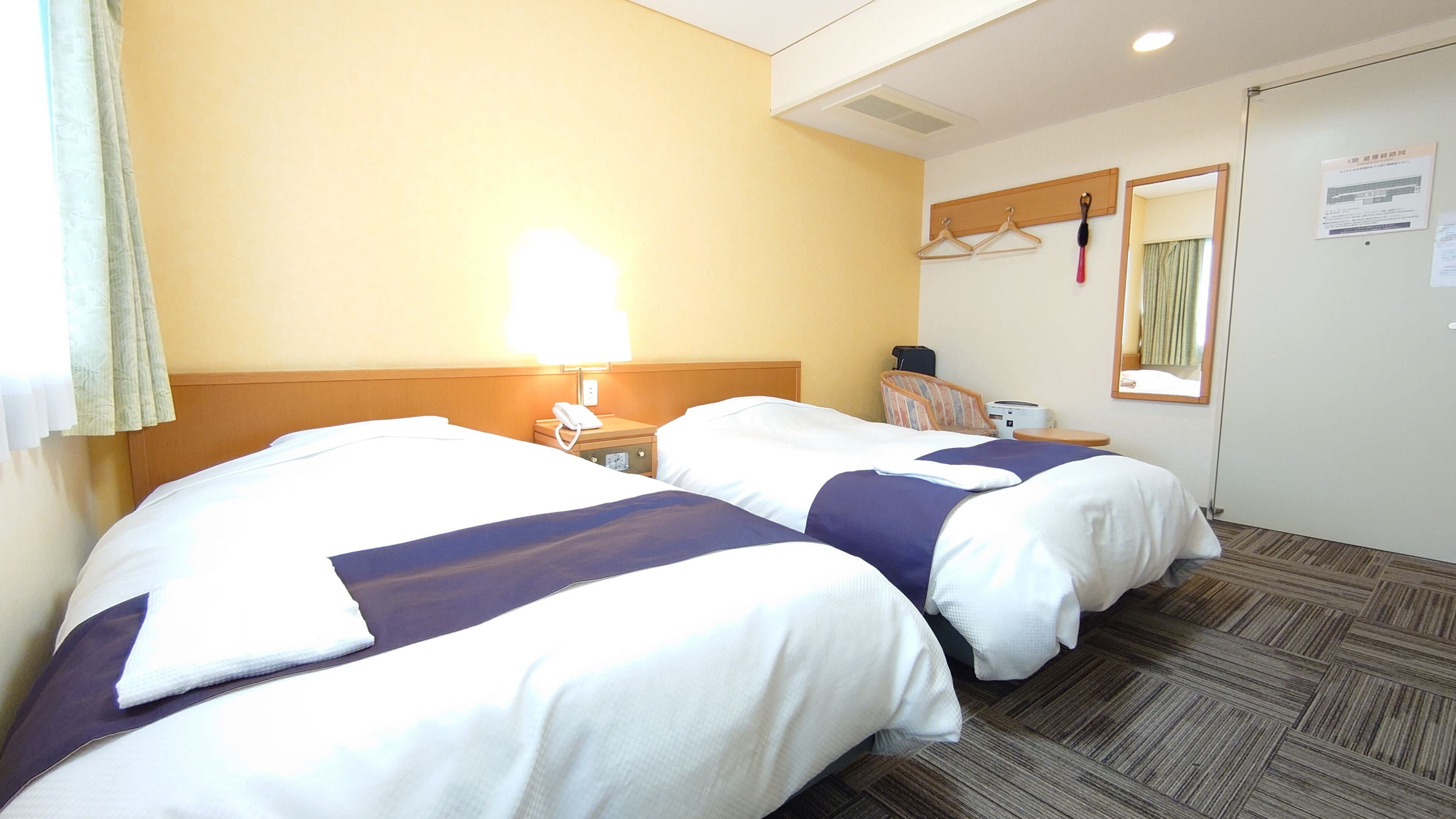 A hotel room with two beds, a bedside table with a lamp, a chair, a mirror, and a coat rack.
