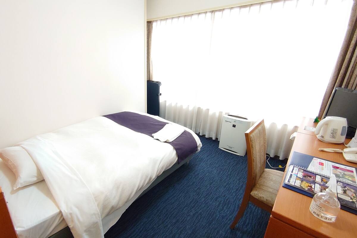 A hotel room with a single bed, a desk with a chair, a radiator, a kettle, and a small table with a bottle.