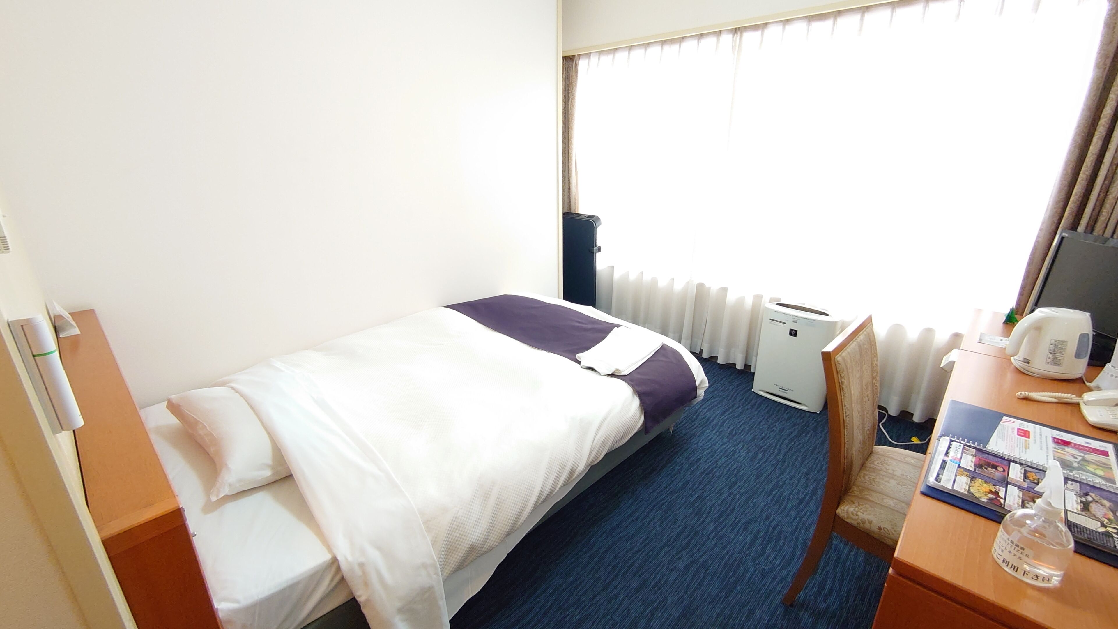 A hotel room with a bed, a desk with a computer, a radiator, and a window with curtains.